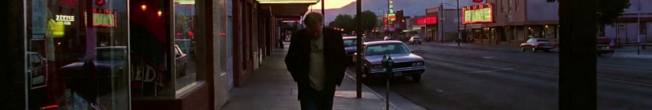 A man with close-cropped grey hair walks east on Central Avenue at dusk and passes out of frame, leaving the neon-lit street empty